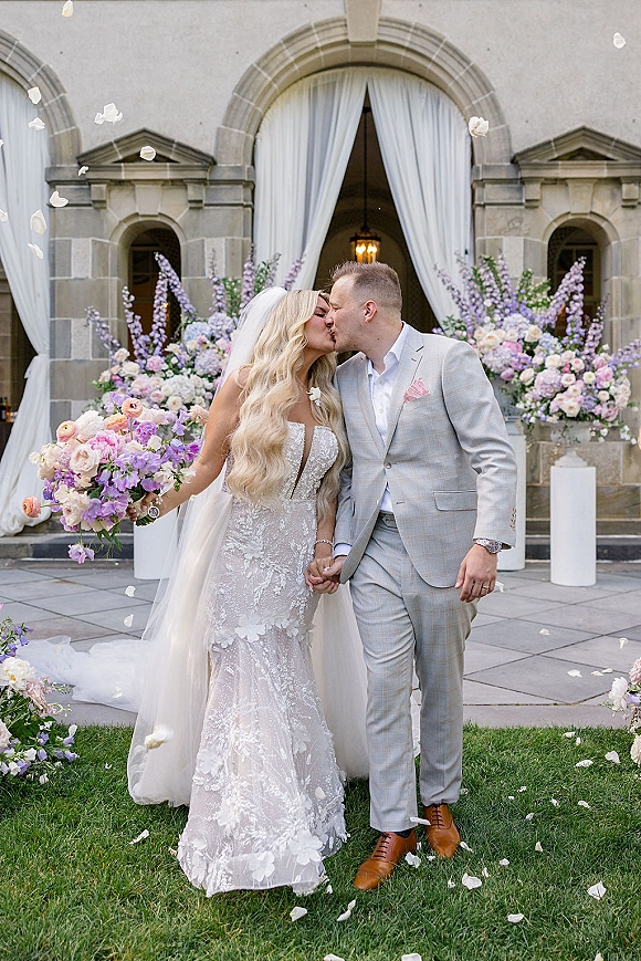 Wedding kiss portrait of bride and groom kissing under a floral arch, bouquet and long veil, petals falling by a stone doorway
