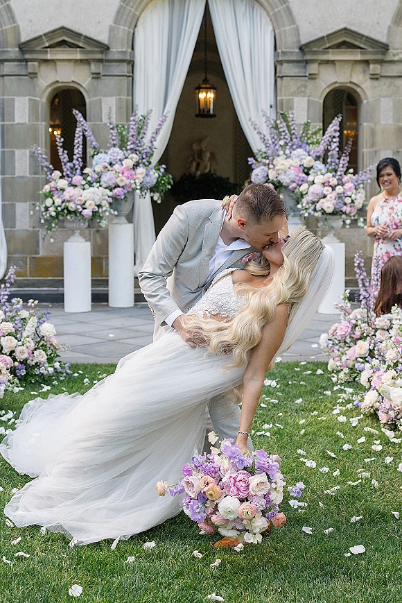 Wedding kiss portrait of groom dipping the bride in a lace dress with veil, pastel bouquet and floral pedestals by a stone archway entrance