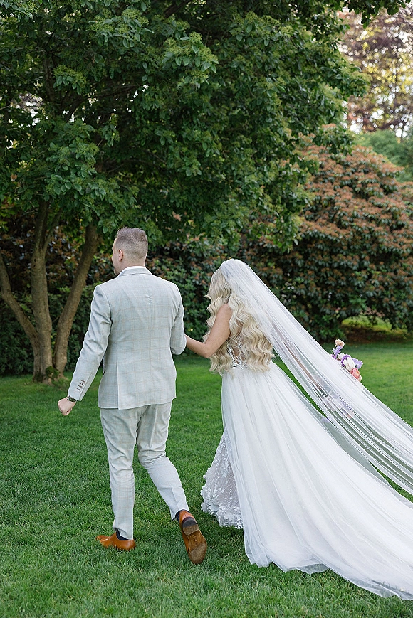 Couple portrait of bride and groom walking away, her long veil trailing over grass, arm in arm on a garden lawn with trees and hedges