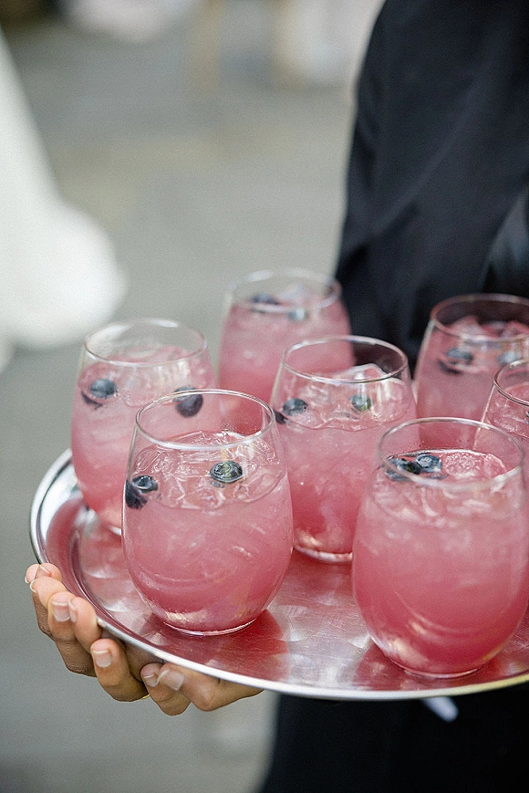 Wedding signature drinks, pink wedding cocktails in stemless glasses with ice and blueberries on a tray held by a tuxedoed server indoors