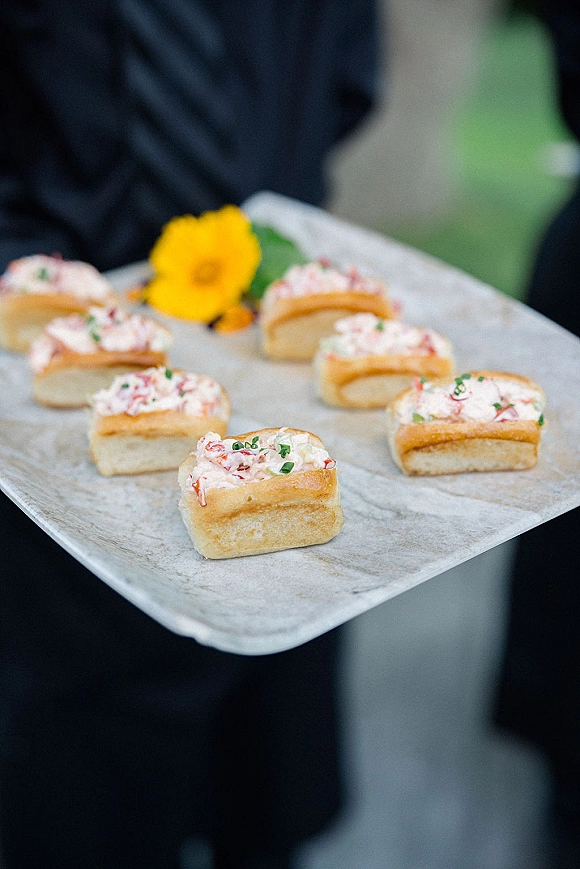 Wedding passed appetizers on a cocktail tray, mini lobster salad sandwiches topped with chives garnish, held by a server amid greenery