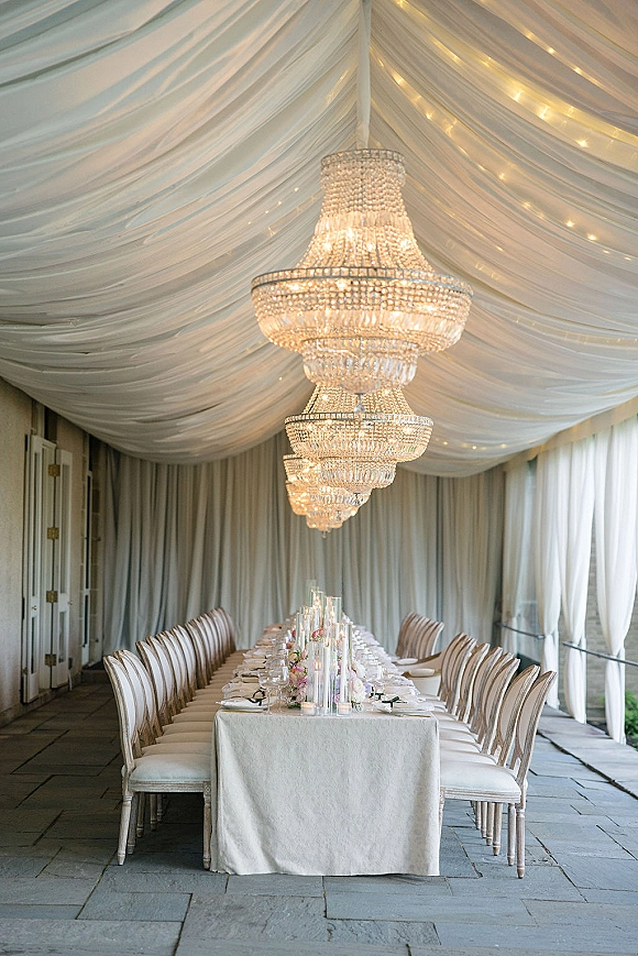 Reception tablescape with long banquet table wedding decor, floating candles and floral centerpiece beneath a crystal chandelier on a covered patio