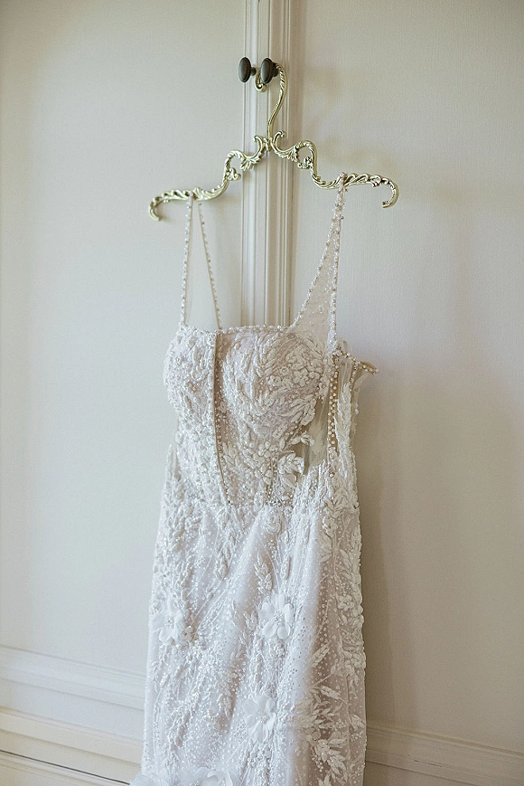 Wedding dress with lace wedding dress detailing, beaded bodice and spaghetti straps on an ornate metal hanger against a white wall and door molding