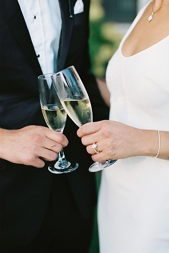 Wedding champagne toast as bride and groom hands clink champagne flutes, rings and jewelry visible against soft outdoor greenery background