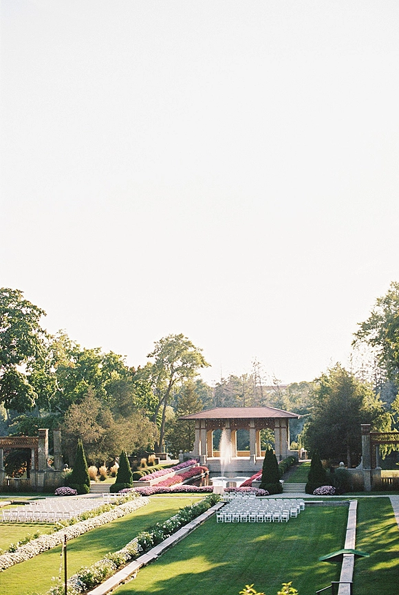 Outdoor ceremony setup with white folding chairs lining a long aisle beside manicured hedges, facing a pavilion near a reflecting pool
