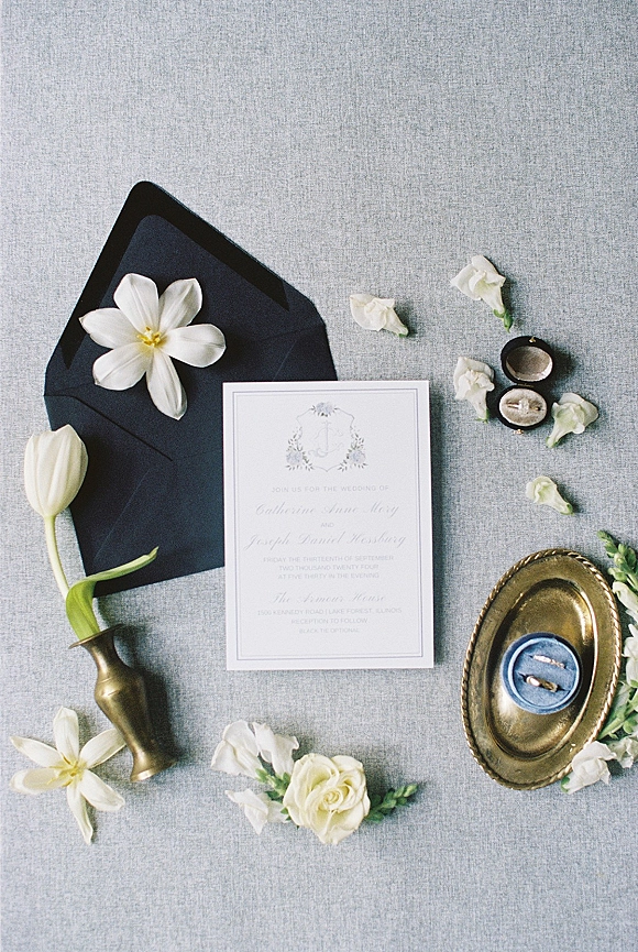 Invitation suite wedding invitation flat lay with black envelope, white flowers, rings and brass tray arranged on gray linen fabric