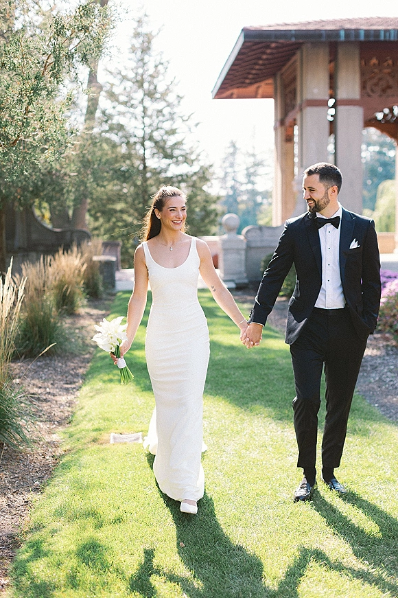 Couple portrait of bride and groom holding hands, her white calla lily bouquet and his black tuxedo, walking on a sunny garden lawn