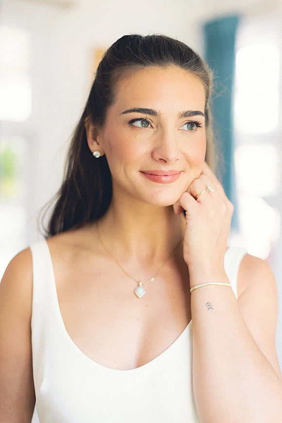 Bridal portrait of a bride getting ready, touching her face in window light, showing white dress neckline, pendant necklace, and ring