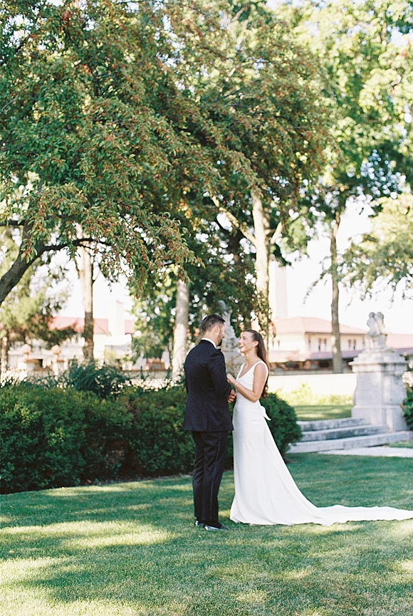 Couple portrait of bride and groom holding hands, her white wedding dress train flowing beside his black tux on a garden lawn near stone steps