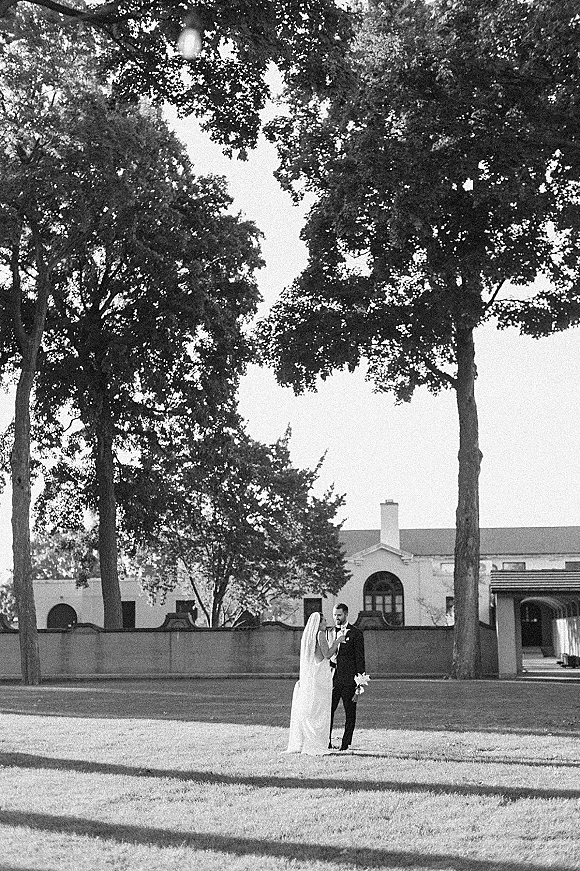 Couple portrait in black and white wedding portrait style, bride in wedding dress with long veil holding bouquet beside groom in tuxedo on lawn near trees
