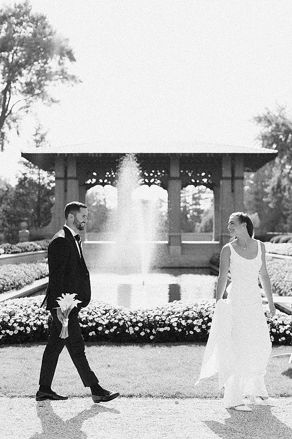 Couple portrait in a black and white wedding photo, bride in sleeveless gown and groom in tuxedo by a garden fountain and reflecting pool