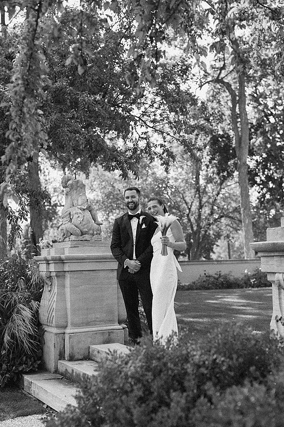 Couple portrait in a black and white wedding portrait style, bride holding calla lily bouquet beside groom in tuxedo on stone steps in a garden