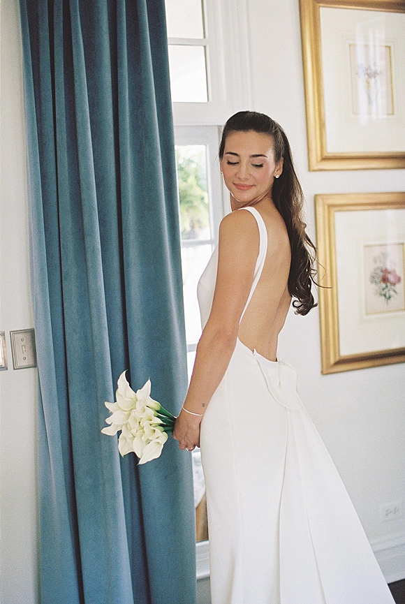 Bridal portrait of a bride in an open back wedding dress holding a calla lily bouquet, looking over her shoulder by a window with blue curtains