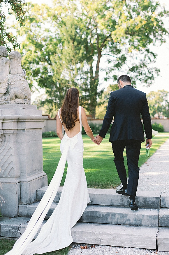 Couple portrait of bride and groom walking away holding hands, her low-back gown with long train, groom holding bouquet by stone steps in a garden lawn