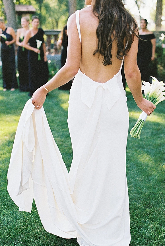 Bride portrait in a backless wedding dress, walking away holding a calla lily bouquet, with trees, fence, and bridesmaids behind.