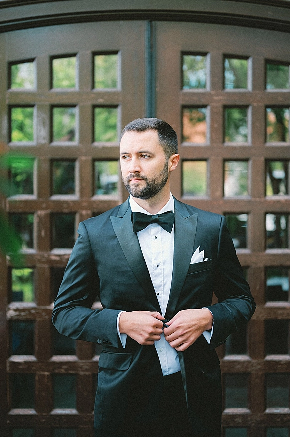 Groom portrait in a black tuxedo, bow tie, and pocket square, standing by wood paneled doors with window panes and greenery outside