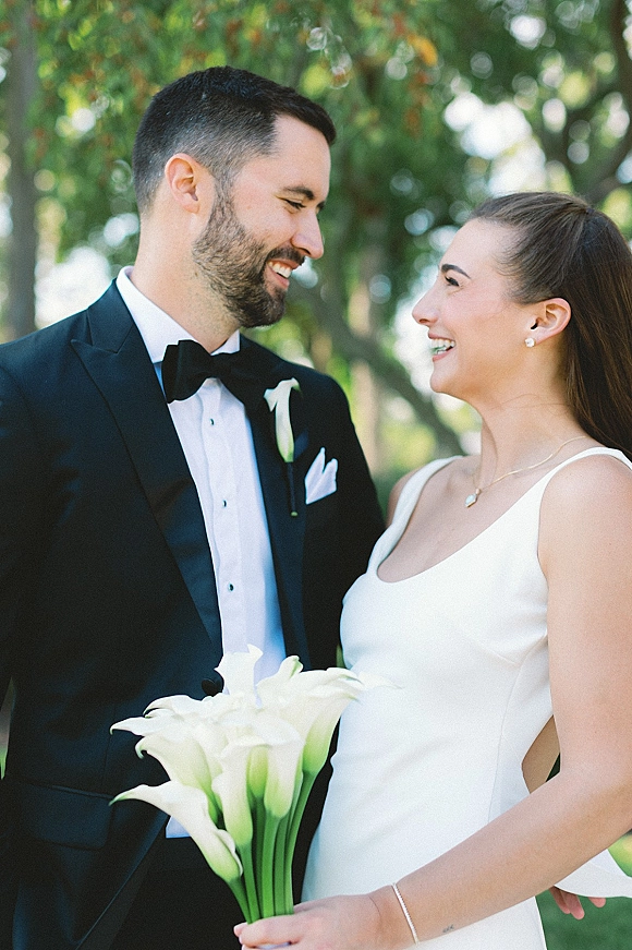 Couple portrait of bride and groom smiling, gazing at each other in sunlight; he wears a black tuxedo, she holds calla lilies.