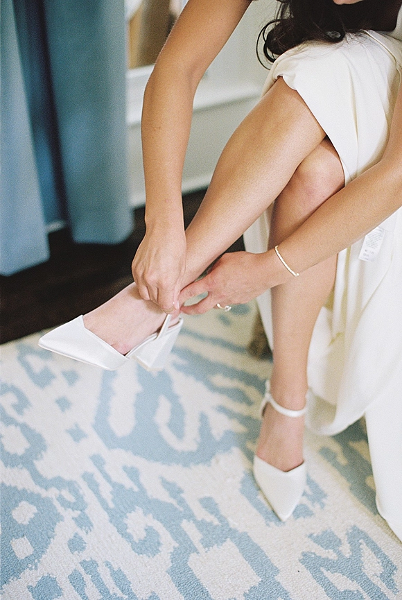 Bridal shoes as the bride fastens white wedding heels with ankle strap while seated in a robe on a patterned rug near curtains
