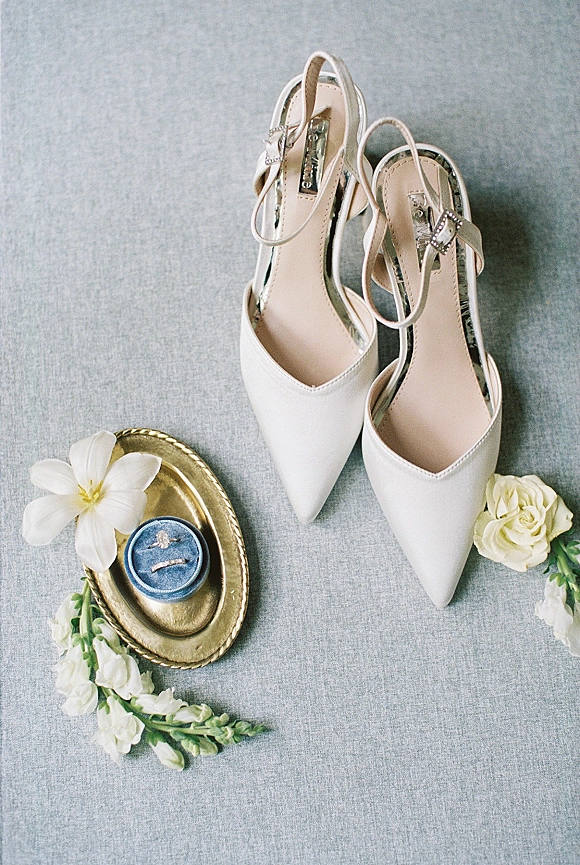 Bridal shoes in a wedding shoe flat lay with white pointed-toe ankle-strap heels, rings, blue velvet box, flowers on gray fabric