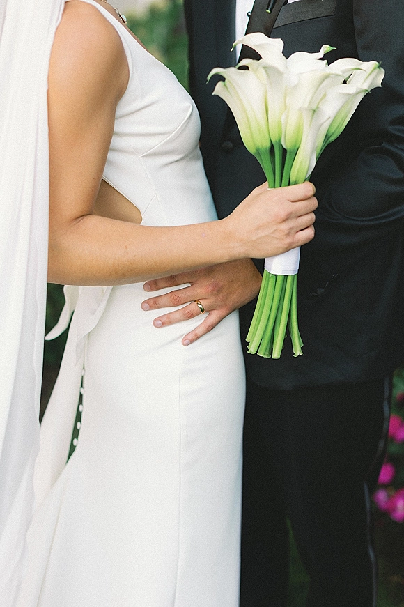 Couple portrait of bride and groom close up, bride holding a white calla lily bouquet, groom’s hand at her waist in a blurred garden