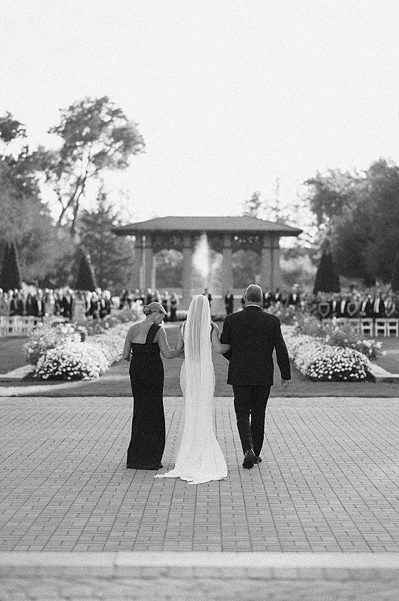 Bride walking down aisle with a long cathedral veil, escorted by parents along a floral-lined brick walkway at an outdoor garden pavilion ceremony