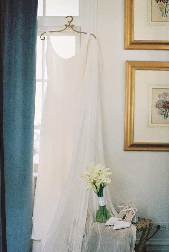 Wedding dress hanging by a window with blue curtain, veil draped over it, and calla lily bouquet, shoes, and ribbon wrap nearby