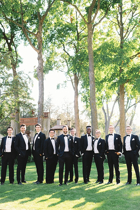 Groomsmen portrait in classic black tuxedos and bow ties, lined up on a green lawn with tall trees and a stone wall behind them