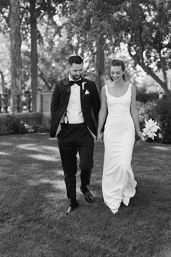 Couple portrait of bride and groom walking hand in hand, she holds a lily bouquet, in dappled sunlight on a garden lawn by a stone balustrade