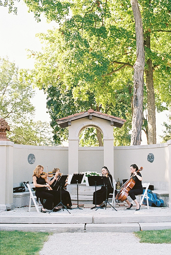 Ceremony musicians with string instruments and sheet music on stands, seated by white folding chairs in an outdoor courtyard with stone archway