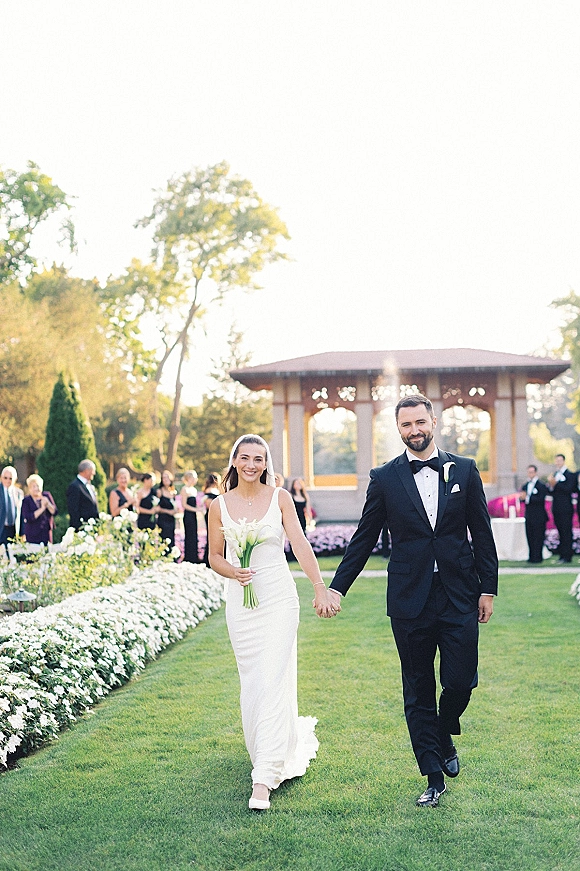 Wedding recessional as bride and groom walking hand in hand, bride holding calla lily bouquet and veil, guests applauding on garden lawn near gazebo