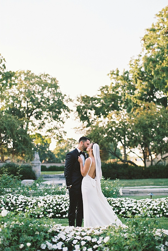 Wedding kiss portrait of bride and groom kissing in a garden, her long veil draped over an open-back dress near a stone fountain