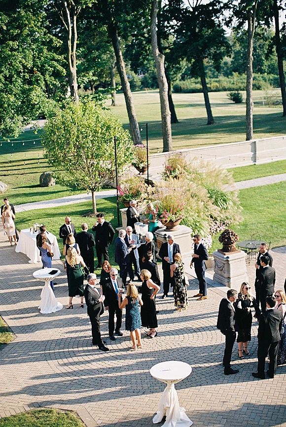 Cocktail hour with wedding cocktail hour guests mingling around high-top tables with white linens and string lights on a stone patio garden