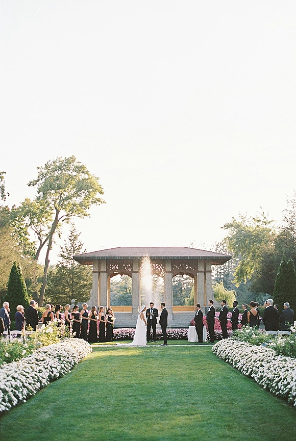 Outdoor wedding ceremony with bride in veil and groom at a garden ceremony setup under a pavilion arch, fountain and trees behind them