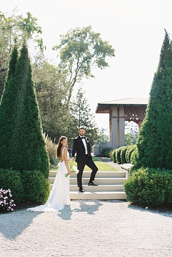 Couple portrait of bride holding bouquet and groom in tuxedo holding hands on stone steps in a manicured garden near a pavilion
