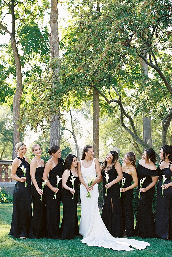Bride with bridesmaids in black dresses holding white calla lily bouquets on a garden lawn by a stone balustrade