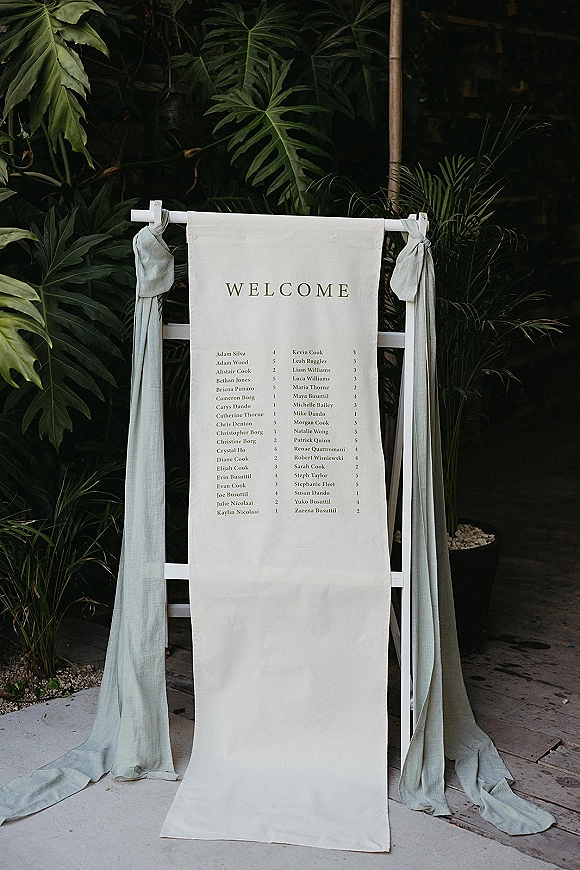 Wedding welcome sign with a fabric welcome banner on a white stand, draped sage fabric, set by tropical greenery on a wooden deck