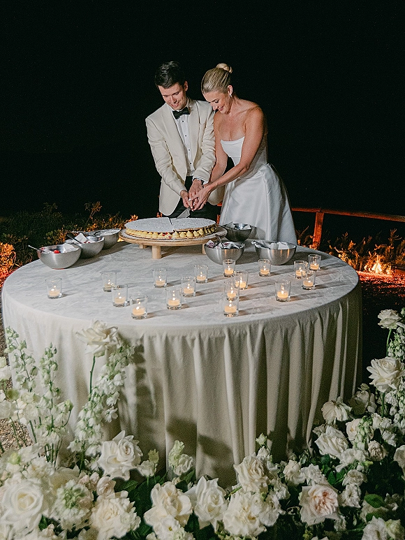 Wedding cake cutting as bride and groom slice an oversized cake on a candlelit table with white roses under a starry outdoor backdrop