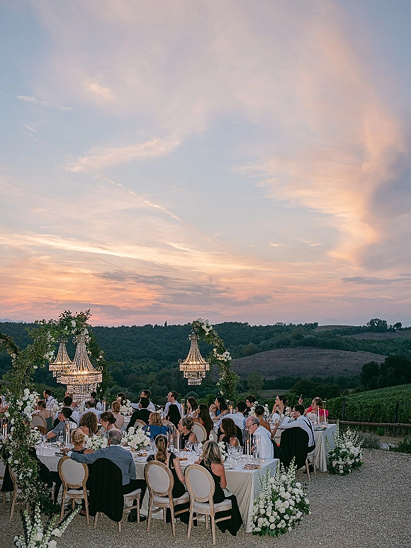Outdoor wedding reception with long banquet tables in white linens, crystal chandeliers, and candlelit florals set against a vineyard sunset