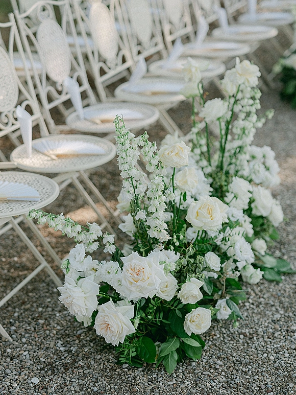 Ceremony aisle florals with white roses, delphinium, and greenery lining a gravel path beside white folding chairs with programs
