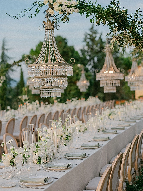 Reception tablescape with a long banquet table wedding setup, white linens, floral garlands, taper candles, and crystal chandeliers under trees