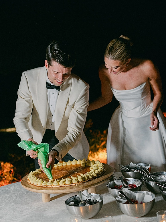 Wedding cake cutting as groom pipes frosting on the cake while bride watches at a candlelit dessert table with berries against a dark night backdrop