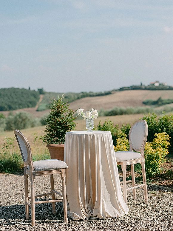 Outdoor cocktail table with ivory tablecloth and crystal vase of white flowers, set with upholstered bar stools on a gravel patio with hills beyond