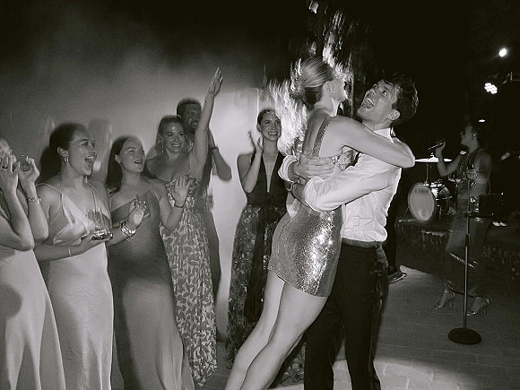Reception dance moment as guests cheer around the wedding dance floor, bride in a sequined mini dress lifted near stage lighting and drum kit