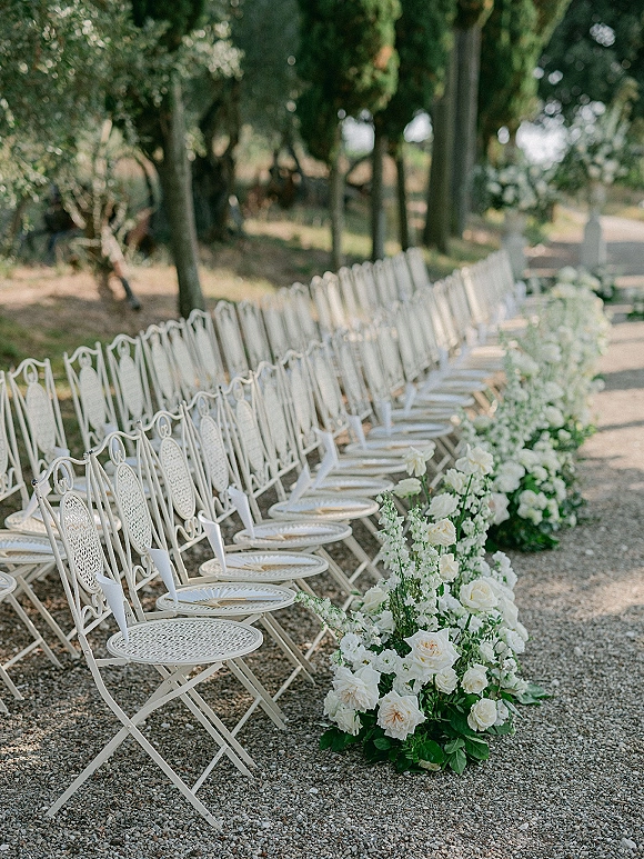 Ceremony aisle decor with outdoor ceremony seating, white wrought iron chairs and white rose greenery arrangements along a gravel garden path