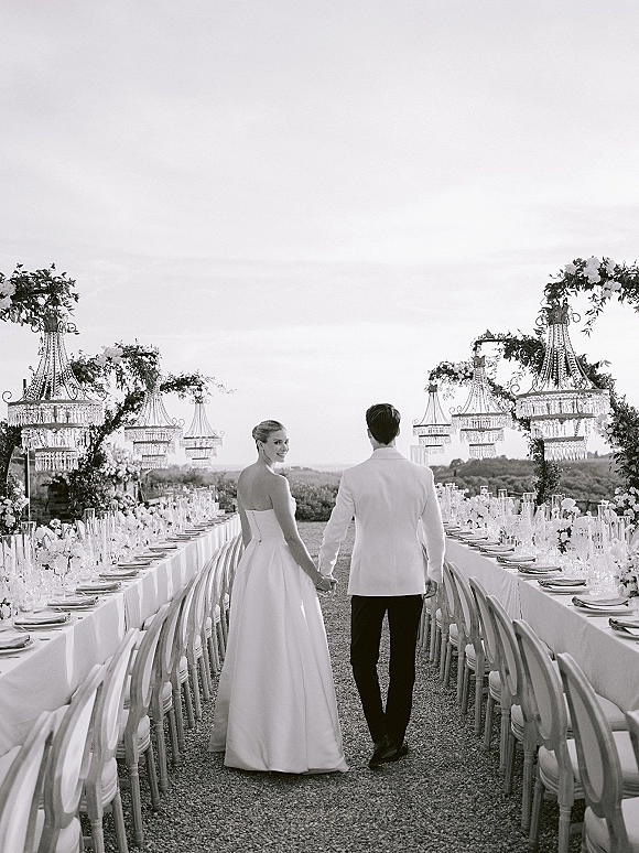 Couple portrait of newlyweds holding hands walking away between long banquet tables with chandeliers and floral garlands under open sky