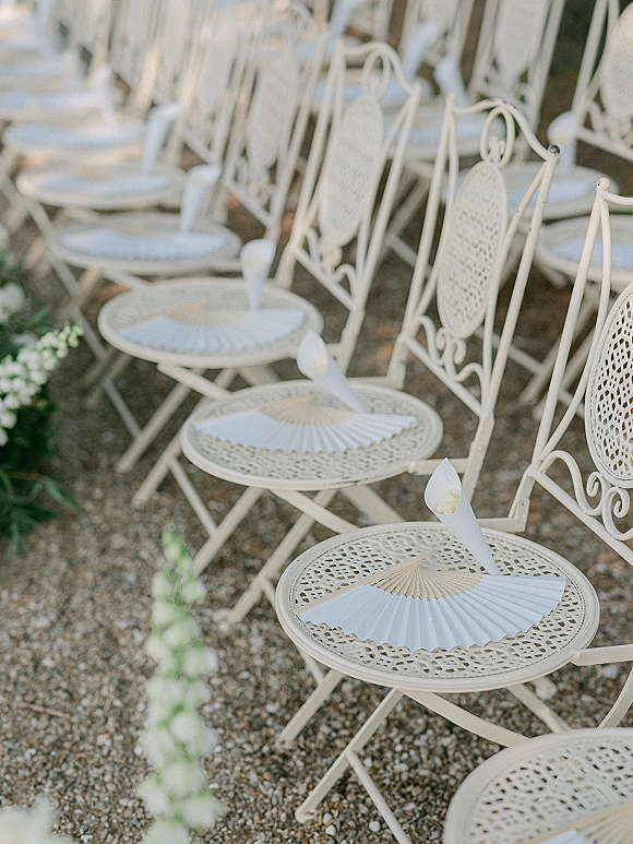 Ceremony seating with outdoor ceremony chairs, white metal scrollwork backs and paper hand fans on a gravel aisle with calla lilies