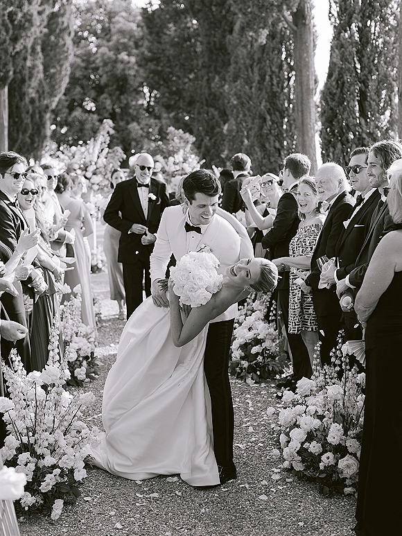 Wedding recessional as groom dips the bride for a kiss, bouquet in hand, guests cheering along a floral-lined garden gravel aisle under tall trees