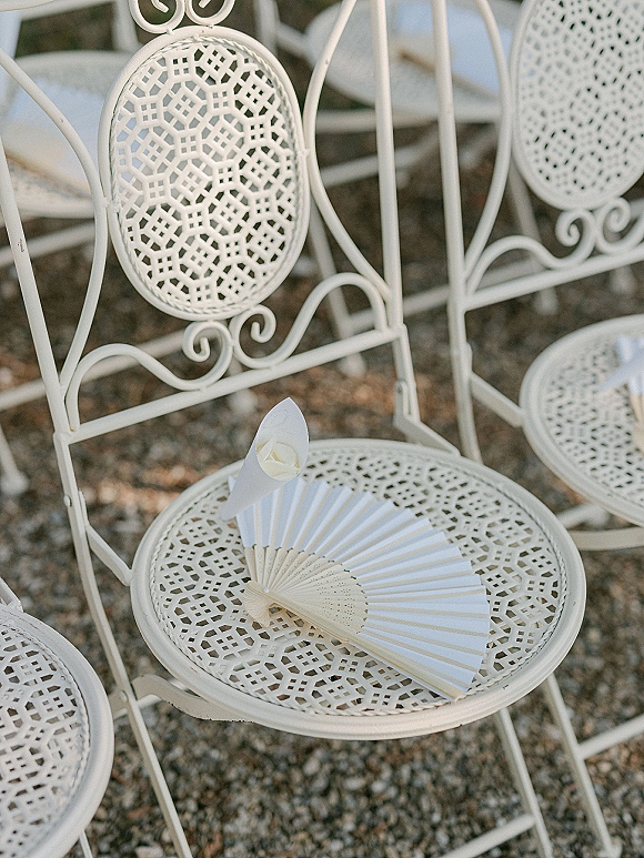 Ceremony chair decor on white metal chairs with a paper fan and flower petal cone on the seat in an outdoor gravel seating area