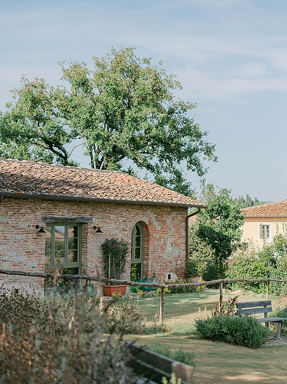 Wedding venue exterior of a rustic wedding venue with a brick farmhouse, terracotta roof, and potted flowers along a garden path under blue sky