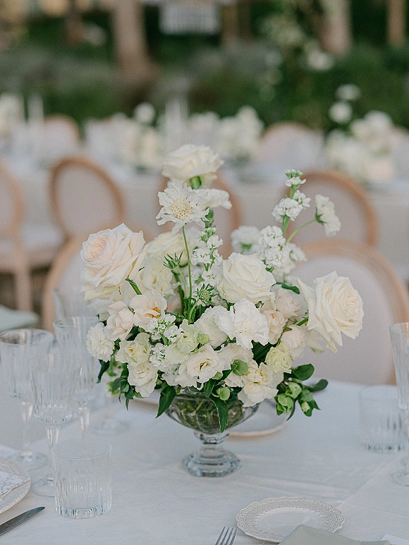 Wedding centerpiece of white roses and greenery in a glass compote vase on a white tablescape with scalloped plates and clear glassware